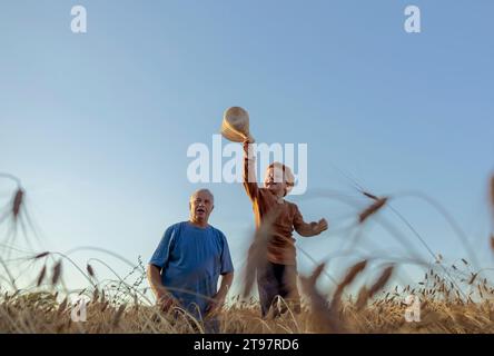 Petit-fils tenant le chapeau de paille avec grand-père dans le champ de blé sous le ciel Banque D'Images