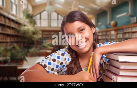 Heureuse jeune fille hispanique avec un crayon et une pile de livres faisant le travail à domicile dans une bibliothèque Banque D'Images