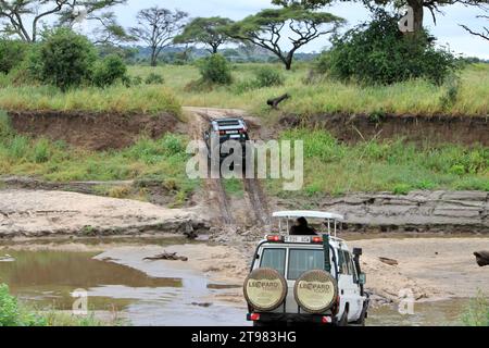 Un convoi de plusieurs véhicules fait son chemin sur une route boueuse sinueuse, passant à travers un paysage rural luxuriant avec une rivière longeant Banque D'Images