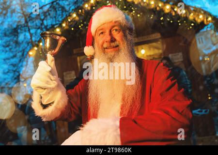 Père Noël tenant une cloche avec une expression joyeuse sur un marché de Noël avec des lumières festives Banque D'Images