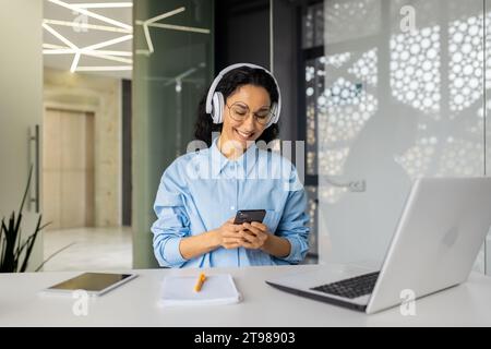 Jeune belle femme au lieu de travail joyeuse écoutant la musique et la radio en ligne, en utilisant l'application sur le téléphone, femme d'affaires reposant sur la pause, chantant le long du travail bien fait bien fait. Banque D'Images