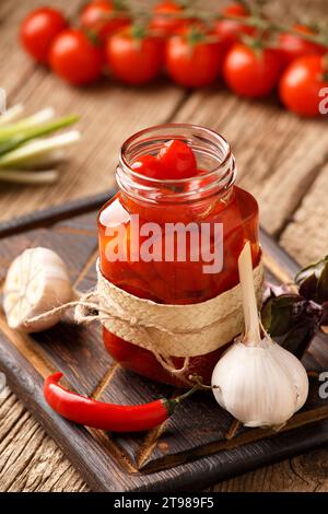 Tomates cerises en conserve dans un bocal en verre ouvert, ail, piments, tomates fraîches, épices et herbes pour marinade sur fond en bois. Banque D'Images