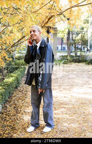 Jeune homme moderne avec un style urbain parlant et souriant avec un téléphone portable en automne dans le parc Banque D'Images
