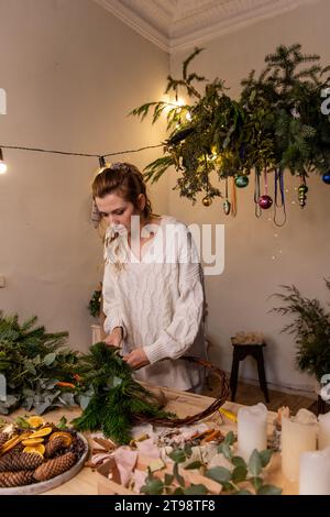 Photo verticale de jeune femme faisant la couronne de Noël à la main à la maison à partir de matériaux naturels. Décoration maison DIY pour les vacances du nouvel an. Blogueur Writ Banque D'Images