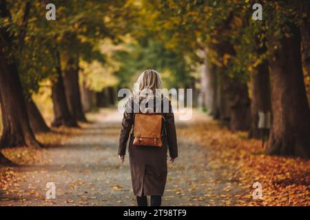 Femme élégante avec trench coat brun et sac à dos marche dans l'allée au parc d'automne. Beauté et mode de vie Banque D'Images