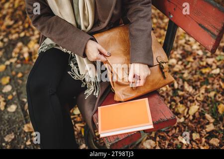 Femme ouvrant sac à main en cuir sur banc dans le parc d'automne. Détendez-vous avec réserver à l'extérieur. Accessoire personnel de mode Banque D'Images