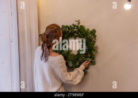 Authentique jeune femme en pull tricoté accroche une couronne de Noël douce et verte en épinette naturelle, pin, branches d'eucalyptus sur le mur de l'appartement. W Banque D'Images