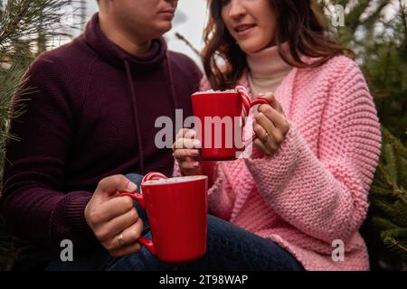 Gros plan des mains d'un jeune couple tenant des tasses rouges avec du cacao chaud, des guimauves, du rouge rayé, de la canne à sucre blanche. L'homme et la femme s'amusent à Christ Banque D'Images