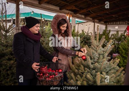 Couple drôle décore les arbres de Noël au marché avec des boules rouges. Jeune homme et femme accrochent des jouets sur le sapin dans le jardin d'arrière-cour, créant confortable, festif Banque D'Images