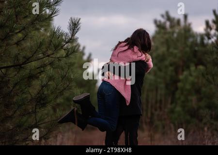 Couple heureux tourne dans la forêt de conifères sur le chemin. Le jeune homme embrasse fermement la femme aux cheveux bouclés dans un pull rose. Fiançailles hivernales, mariage d'automne. Proposa Banque D'Images