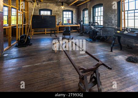 Un rail en cadre métallique rouillé repose sur des tréteaux en bois au premier plan de la salle expérimentale reconstruite de la Ford Piquette Plant à Detroit, Michigan, États-Unis, avec des outils mécaniques et une chaise à bascule devant un tableau noir derrière elle. Dans cette pièce du troisième étage étroitement surveillée, Henry Ford et une petite équipe de confiance ont développé la Ford Model T sur environ un an, à partir de janvier 1907. Construite en 1904, la Ford Piquette Plant est considérée comme le plus ancien bâtiment survivant initialement construit pour la production automobile et qui reste ouvert au public. Banque D'Images