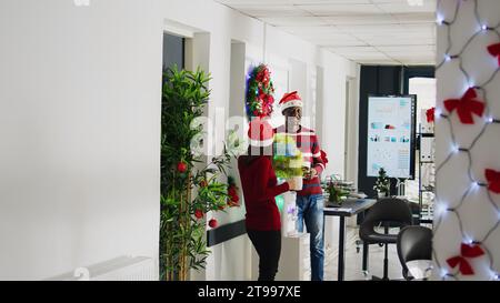 Les employés dans le bureau décoré de Noël appréciant une tasse de café par le refroidisseur d'eau, riant ensemble. Heureux membres du personnel BIPOC portant des chapeaux de Père Noël prenant pause dans l'espace de travail multiracial Banque D'Images