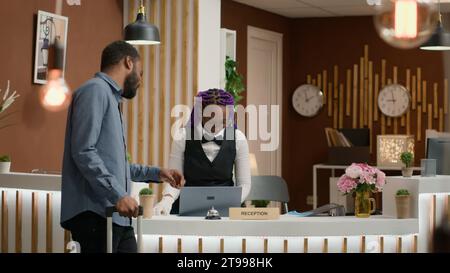 La personne appelle pour le service et la carte de chambre à la réception en utilisant la cloche de la réception pour parler à la réceptionniste dans le hall. Client afro-américain avec sacs de trolley recevant la clé d'accès pour l'hébergement. Banque D'Images