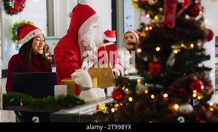 Travailleurs multiethniques recevant des cadeaux d'un collègue prétendant être le Père Noël dans un bureau décoré de Noël. Employé portant le costume surprenant le personnel de l'entreprise avec des cadeaux pendant la saison des vacances d'hiver Banque D'Images