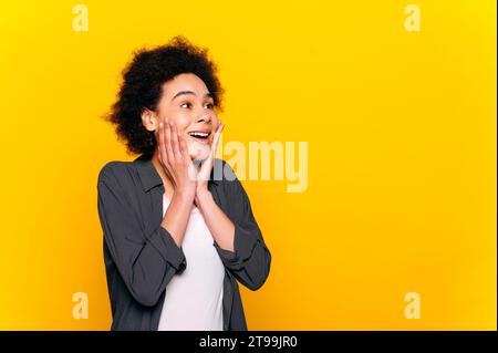 Excité surpris aux cheveux frisés afro-américains élégant femme positive, regarde dans l'étonnement sur le côté à l'espace vide, se tient sur fond jaune isolé, souriant Banque D'Images