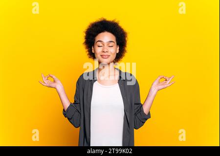 Photo d'une belle femme calme afro-américaine ou brésilienne aux cheveux bouclés dans une chemise décontractée, méditant tout en se tenant debout avec les yeux fermés sur fond jaune isolé. Santé mentale, harmonie, détente Banque D'Images