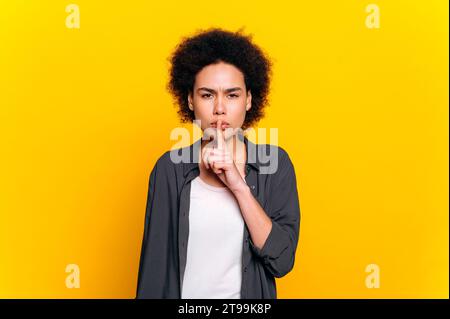 Photo d'une femme afro-américaine, en tenue décontractée, montrant le signe shh avec l'index touchant ses lèvres, arrêter de parler, regarder la caméra, se tenir debout sur fond jaune isolé. Ne le dis pas. Secret, mystère Banque D'Images