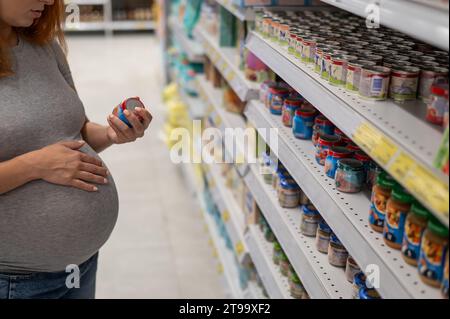 La femme enceinte caucasienne choisit la nourriture pour bébé dans le magasin. Pots avec purée de légumes. Banque D'Images