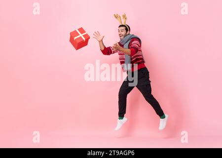 Portrait en longueur d'un homme portant un chandail de Noël et un bandeau fantaisie de renne attrapant la boîte-cadeau en fond isolé de couleur rose Banque D'Images