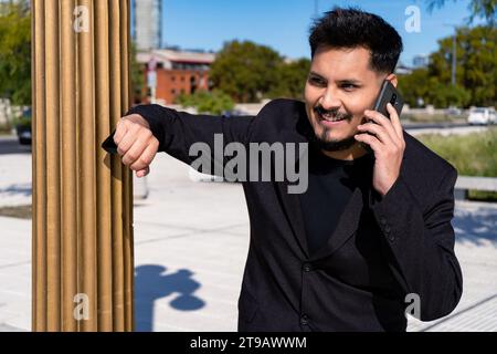 Gros plan d'un jeune homme latino souriant parlant sur son téléphone portable ou son smartphone sur une place publique. Banque D'Images