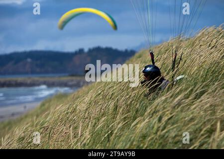 Parapente féminine assise dans de hautes herbes sur une dune au-dessus d'une plage tandis qu'un autre parapente vole en arrière-plan. Banque D'Images