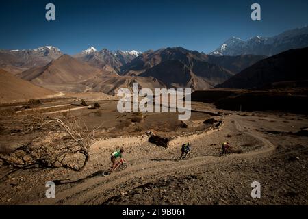 Trois hommes montent en vélo de montagne sur un chemin de terre avec d'énormes montagnes et une petite ferme. Banque D'Images