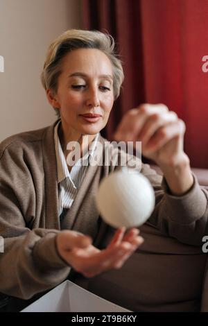 Gros plan de portrait vertical d'une femme d'âge moyen tenant et regardant la boule de Noël avec une expression heureuse. La femelle élégante décore l'arbre de Noël pour Banque D'Images