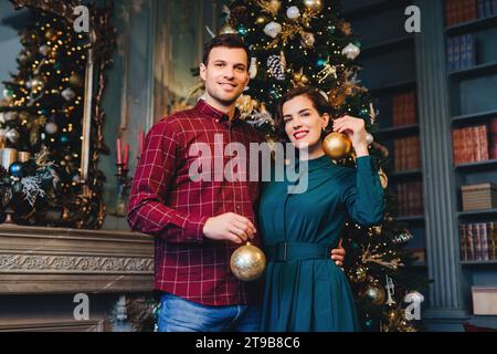 Couple en tenue festive tenant des ornements de Noël dorés, debout près d'un arbre richement décoré dans une pièce élégante. Banque D'Images