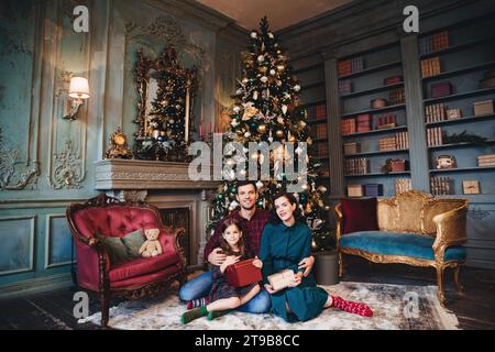 Famille avec un jeune enfant assis dans une chambre de style vintage avec un sapin de Noël richement décoré, tenant des cadeaux Banque D'Images