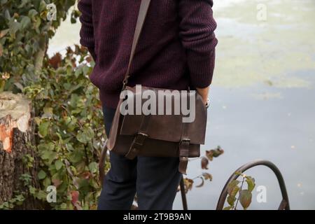 Homme avec sac en cuir sur fond de nature à l'extérieur. Homme décontracté portant un chandail tricoté et un sac pour ordinateur portable. Tenues décontractées pour hommes avec accessoires. Style Banque D'Images