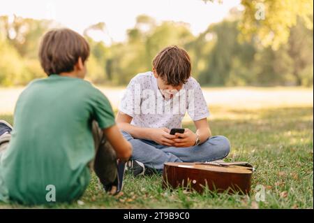 Deux frères passent du temps assis sur l'herbe dans le parc tout en parlant, en jouant du téléphone et de la guitare. Banque D'Images