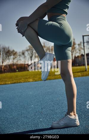 vue partielle de sportswoman flexible s'exerçant en vêtements d'activité serrés à l'extérieur, étirement de la jambe Banque D'Images
