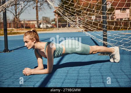 jolie jeune sportswoman avec queue de cheval s'exerçant en vêtements d'activité près du filet à l'extérieur, faisant planche Banque D'Images