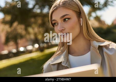 portrait de jolie jeune femme dans des écouteurs sans fil et manteau beige trench regardant loin dans le parc Banque D'Images