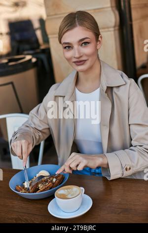 heureuse jeune femme en trench coat profitant de ses gaufres belges avec de la crème glacée à côté d'une tasse de café Banque D'Images