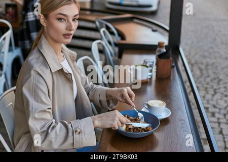 jolie jeune femme en trench coat mangeant ses gaufres belges avec de la crème glacée à côté d'une tasse de café Banque D'Images