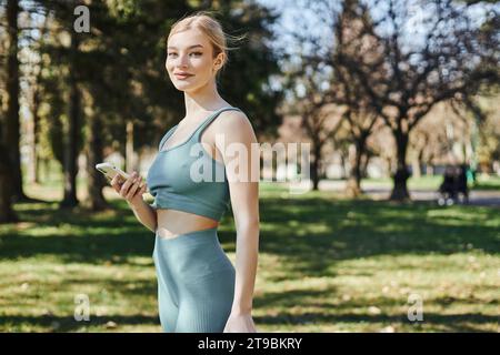 heureuse sportswoman en tenue active tenant le smartphone et regardant l'appareil photo tout en se tenant debout dans le parc Banque D'Images