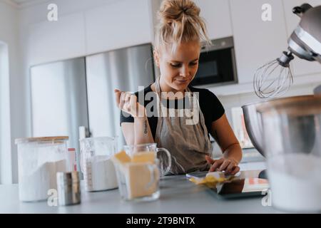 Jeune femme vérifiant le téléphone cellulaire dans la cuisine tout en faisant cuire Banque D'Images