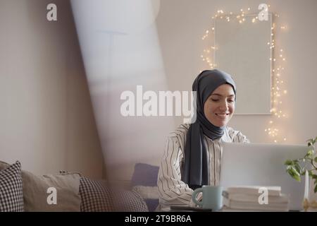 Femme pigiste souriante portant le hijab tout en travaillant sur ordinateur portable dans le salon à la maison Banque D'Images