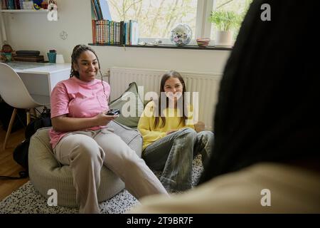 Jeune fille souriante tenant le contrôleur de jeu tout en parlant avec des amies féminines à la maison Banque D'Images