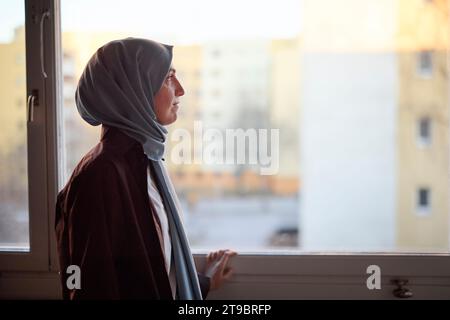 Femme réfléchie en foulard regardant par la fenêtre Banque D'Images