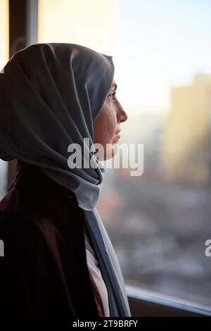 Femme réfléchie en foulard regardant par la fenêtre Banque D'Images