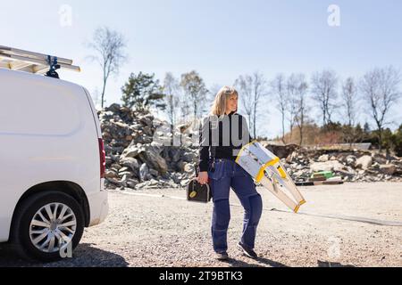 Femme électricienne souriante portant l'échelle et la boîte à outils debout sur la route par van Banque D'Images