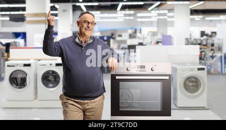 Homme mûr souriant appuyé sur un four et pointant vers le haut à l'intérieur d'un magasin pour les appareils ménagers Banque D'Images