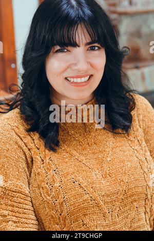 portrait vertical d'une jeune femme heureuse, aux cheveux noirs, mignonne, latine habillée de brun, assise regardant la caméra et souriant Banque D'Images