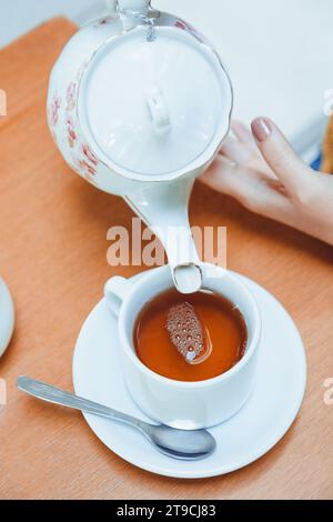 Image verticale et vue de dessus des mains féminines d'une femme caucasienne méconnaissable, versant du thé dans une tasse de porcelaine sur la table sur une table de café Banque D'Images