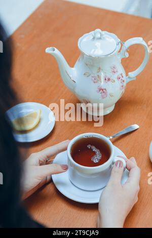 Gros plan vue aérienne d'une femme méconnaissable assise buvant du thé tenant une tasse en porcelaine sur une assiette sur la table. espace de copie. Banque D'Images