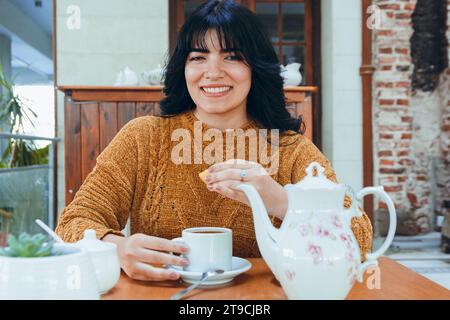 Vue de face de la jeune femme vénézuélienne latine, heureuse souriante et regardant la caméra tout en préparant et pressant le citron de son thé assis dans le restaurant Banque D'Images