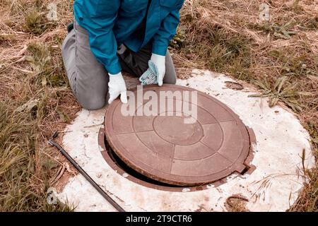 Un travailleur soulève le couvercle de regard sur un puits pour inspection et entretien. Travaux de plomberie en milieu rural. Banque D'Images