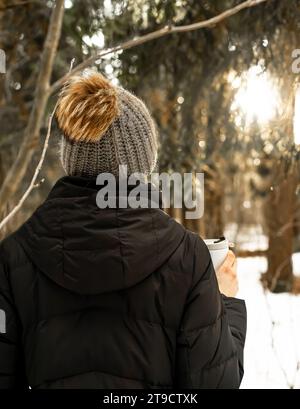 Femme en bonnet en laine tricotée, bonnet avec pompom, tenant le mug thermo dans la nature hivernale Banque D'Images